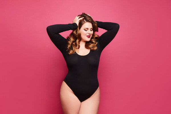 A female plus-size model with bright makeup and trendy hairstyle in black bodysuits over pink background, isolated.