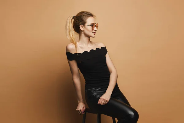 Beautiful and sexy blonde model girl with perfect sporty body and makeup in black leather pants, in black t-shirt and stylish sunglasses sits on stool and and posing at orange background — Stock Photo, Image