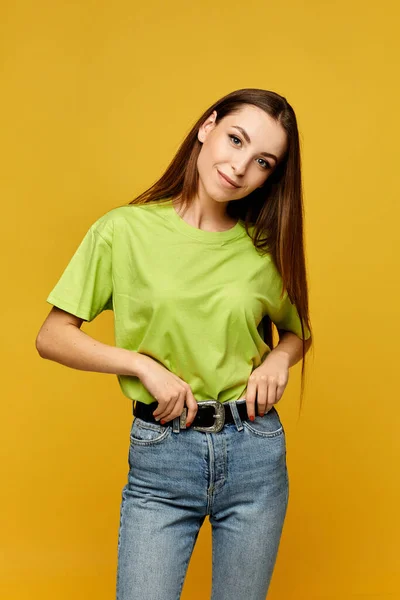 Jovem estudante menina em uma camiseta amarela em branco e jeans posando no fundo amarelo, isolado — Fotografia de Stock
