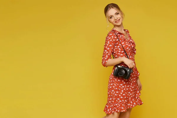 A young woman in summer dress posing with a photo camera on the yellow background, isolated. Traveler concept — Stock Photo, Image