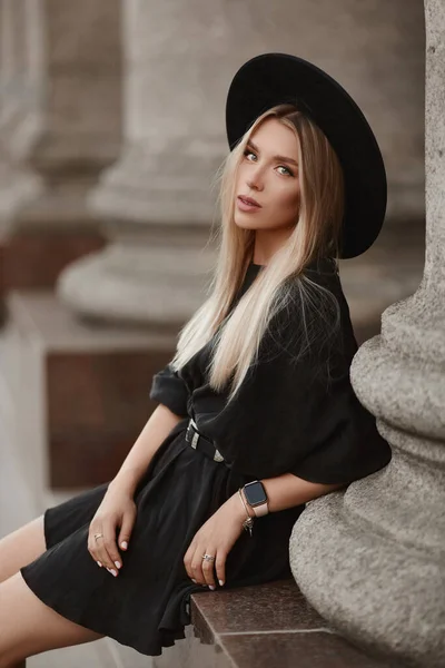 Молодая модель девушка с светлыми волосами в модной шляпе и черном платье позирует на открытом воздухе в городе — стоковое фото
