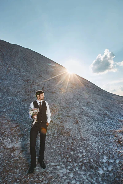 Handsome groom in a stylish suit holding a bridal bouquet and waiting for the bride outdoors over the mountains — Stock Photo, Image