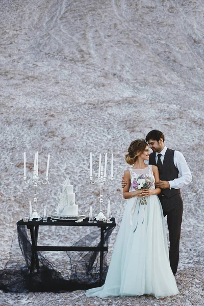 Junge Braut im modischen Kleid und hübscher Bräutigam im eleganten Anzug stehen neben einer luxuriösen Hochzeitstorte im Freien — Stockfoto