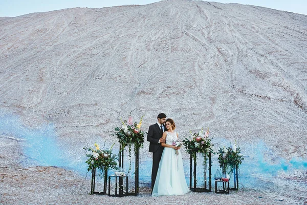 Novia joven en vestido de moda y novio guapo en traje elegante posando en el lugar de la ceremonia de boda decorado con flores al aire libre —  Fotos de Stock