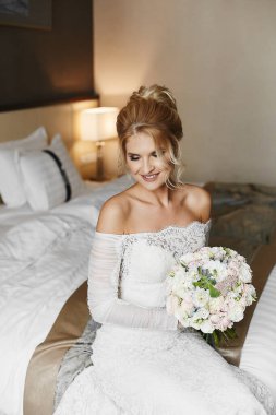 Sarı saçlı ve dantel elbiseli genç bir gelin şık bir iç mekânda yatağında bir buket çiçekle oturuyor.