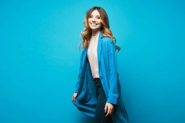 Mavi arkaplanda uzun mavi bir hırkayla gülümseyen genç bir model.