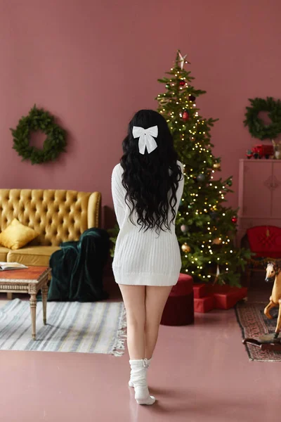 크리스마스 트리 근처에 있는 아늑 한 스웨터를 입은 한 모델 소녀가 신년 휴일을 위해 장식 한 거실 내부에 있는 크리스마스 트리 근처에 포즈를 취하고 있다. — 스톡 사진