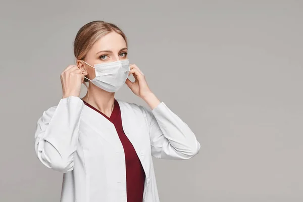 Doctora joven con una capa médica que se pone una cubierta protectora para la cara, aislada en un fondo blanco. Concepto de asistencia sanitaria y protección contra virus. — Foto de Stock