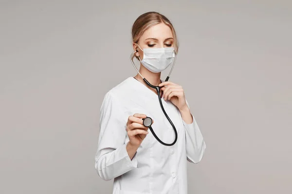 Doctora adulta en uniforme médico y cubierta facial protectora posando con un estetoscopio en el fondo gris, aislada. Concepto de salud y medicina — Foto de Stock