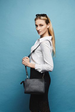 Mavi arka planda beyaz bluz ve siyah dar etek giyen ince vücutlu genç bir bayan.