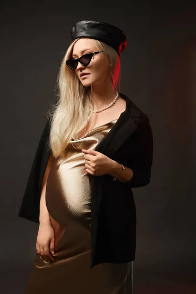 Fashionable young pregnant woman in a gold dress, sunglasses and trendy hat at the dark background. Concept of style for pregnancy.
