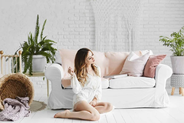 Beyaz saten pijamalı güzel bir kadın beyaz bir iç mekânda kanepenin yanında oturuyor..