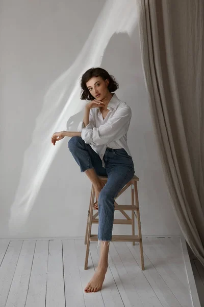 Mulher de meia idade com corpo magro em uma camisa branca e jeans azul olhando na câmera e posando em um banquinho dentro de casa — Fotografia de Stock
