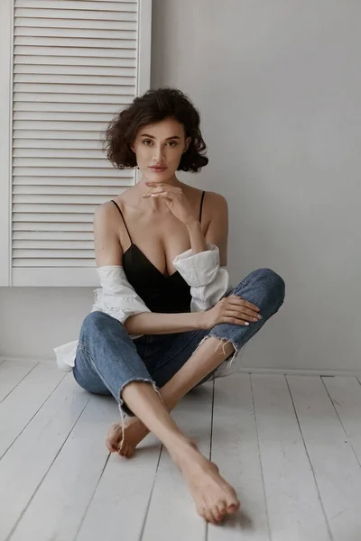 Шикарная девушка-модель с стройным телом в синих джинсах и соблазнительном топе сидит на полу в помещении — стоковое фото