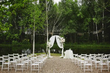 Açık hava düğün töreni seti. Düğün kemeri taze çiçekler ve boş sandalyelerle süslenmiş açık havada küçük göl kenarına yerleştirilmiş..