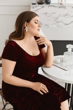 Parlak makyajlı ve son moda küpeli genç şişman kadın oturma odasında kadife bir elbise giyiyor. XXL moda kavramı