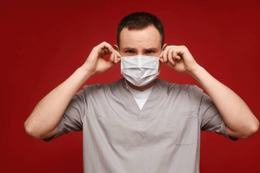 Tıbbi üniformalı genç bir doktor kırmızı arka planda koruyucu bir maske takıyor.