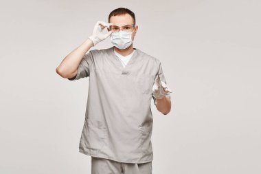 Tıbbi üniformalı, koruyucu gözlüklü ve tıbbi koruyucu maskeli genç adam cerrah makasını tutuyor ve beyaz arka planda izole bir şekilde poz veriyor. Sağlık ve Acil Durum Konsepti