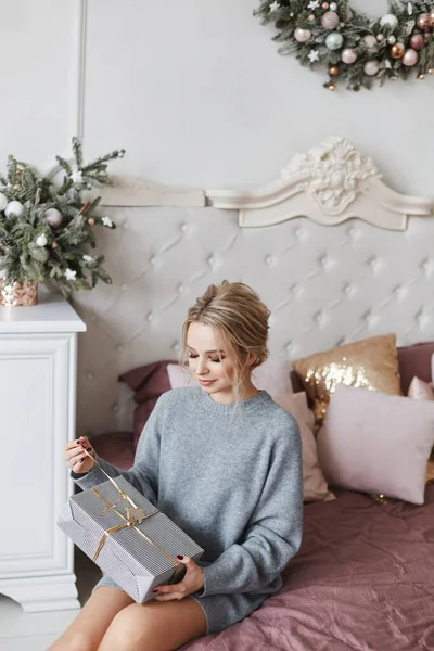 Modische jonge vrouw uitpakken geschenkdoos op het bed in het versierde interieur in de Nieuwjaar ochtend — Stockfoto