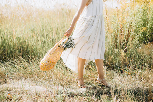 Woman in natural clothes with an eco mesh string bag vegetables and wildflowers walks along a path on the field