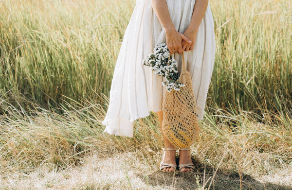 Woman in natural clothes with an eco mesh string bag vegetables and wildflowers walks along a path on the field