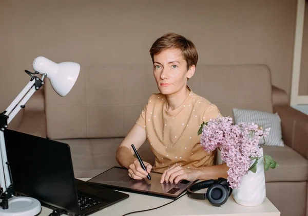 happy woman draws on graphic tablet in bright office with laptop