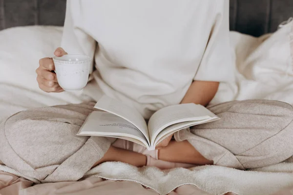 girl in pajamas reads a book and holds a cup of tea inside bedroom