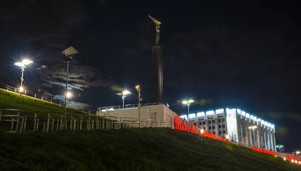 Monumen kemuliaan dalam bentuk prasasti dengan seorang pria dengan sayap di tangan dan bangunan pemerintah di malam hari di Samara Rusia. 26 Juni 2018 — Stok Foto