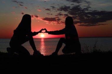 Deniz kıyısındaki iki kızın siluetleri. İki kız bir kalp gösterir. Günbatımında aşk.