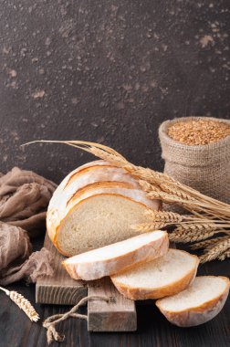 Buğday taneleri ve buğday spikelets, bir çanta ile kahverengi ahşap bir masa üzerinde bir kesme tahtası üzerinde dilimlenmiş beyaz buğday ekmek gönderiyor. Uzay kopyalayın. Önden görünüm. Yakın çekim. Eve ekmek. Ev yapımı. 