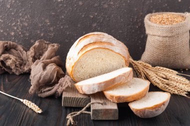 Buğday taneleri ve buğday spikelets, bir çanta ile kahverengi ahşap bir masa üzerinde bir kesme tahtası üzerinde dilimlenmiş beyaz buğday ekmek gönderiyor. Uzay kopyalayın. Önden görünüm. Yakın çekim. Eve ekmek. Ev yapımı. 