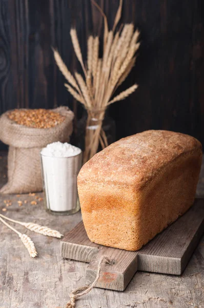 Kepekli ekmek. Mayasız kepekli ekmek unu, buğday taneleri, spikelets ahşap bir kahverengi masa üzerinde bir kesme tahtası üzerinde. Bir somun (tuğla) ekmek. Koyu arka plan. Ev yapımı. 