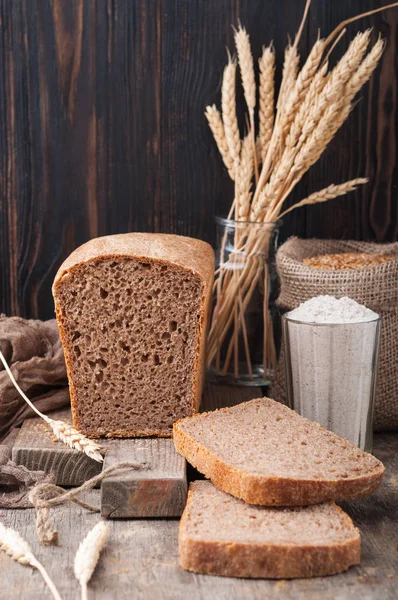 Kepekli ekmek. Mayasız kepekli ekmek unu, buğday taneleri, spikelets ahşap bir kahverengi masa üzerinde bir kesme tahtası üzerinde. Bir somun ekmek parçalar halinde kesilerek. Uzay kopyalayın. Koyu arka plan. Ev yapımı. 