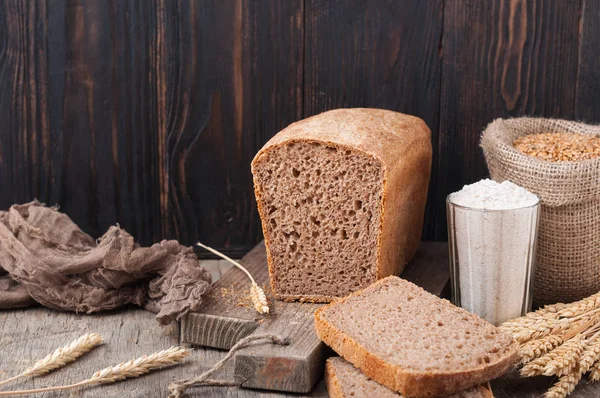 Kepekli ekmek. Mayasız kepekli ekmek unu, buğday taneleri, spikelets ahşap bir kahverengi masa üzerinde bir kesme tahtası üzerinde. Bir somun ekmek parçalar halinde kesilerek. Uzay kopyalayın. Koyu arka plan. Ev yapımı. 