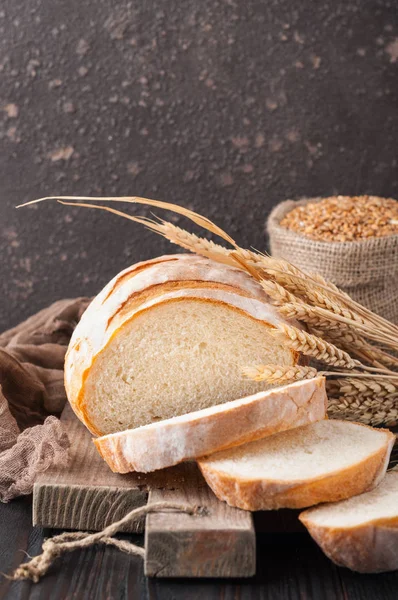 Buğday taneleri ve buğday spikelets, bir çanta ile kahverengi ahşap bir masa üzerinde bir kesme tahtası üzerinde dilimlenmiş beyaz buğday ekmek gönderiyor. Uzay kopyalayın. Önden görünüm. Yakın çekim. Eve ekmek. Ev yapımı. 