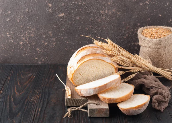 Buğday taneleri ve buğday spikelets, bir çanta ile kahverengi ahşap bir masa üzerinde bir kesme tahtası üzerinde dilimlenmiş beyaz buğday ekmek gönderiyor. Uzay kopyalayın. Önden görünüm. Yakın çekim. Eve ekmek. Ev yapımı. 