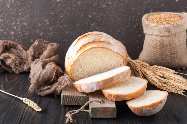 Buğday taneleri ve buğday spikelets, bir çanta ile kahverengi ahşap bir masa üzerinde bir kesme tahtası üzerinde dilimlenmiş beyaz buğday ekmek gönderiyor. Uzay kopyalayın. Önden görünüm. Yakın çekim. Eve ekmek. Ev yapımı. 