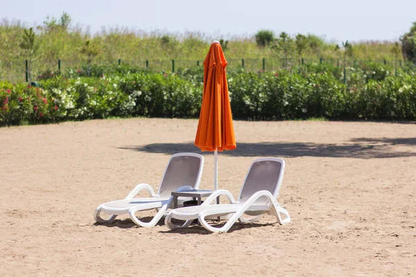 Two chaise lounges stand on the sand with a closed umbrella