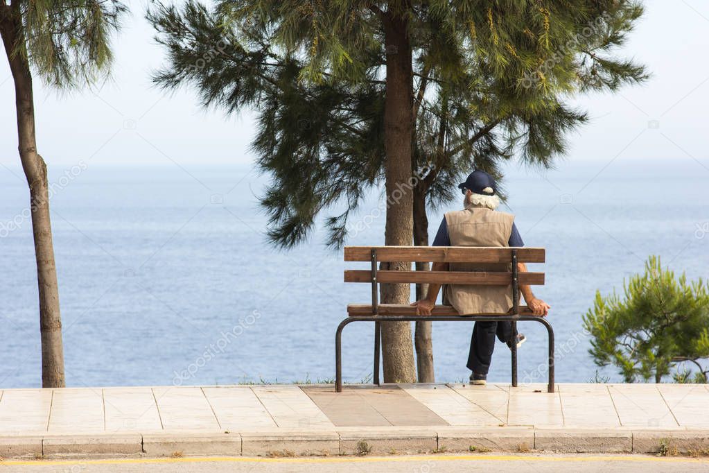 An elderly man sits on a bench by the sea and looks into the distance
