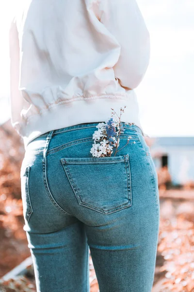 The girl is standing with her back in blue jeans, flowers in her pocket.