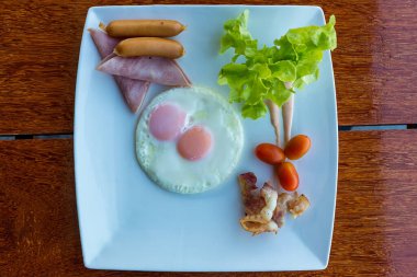 Amerikan kahvaltısı sosisi, yumurta, jambon, pastırma ve sebzeler beyaz tabakta. Kızarmış yumurta ve sosisli geleneksel kahvaltı. Tahta masada kızarmış yumurta, sosis ve jambon..