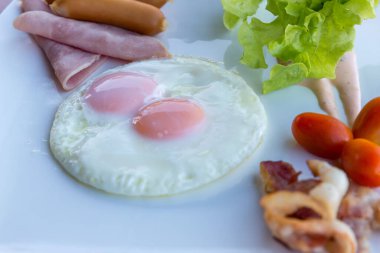 Amerikan kahvaltısı sosisi, yumurta, jambon, pastırma ve sebzeler beyaz tabakta. Kızarmış yumurta ve sosisli geleneksel kahvaltı. Tahta masada kızarmış yumurta, sosis ve jambon..