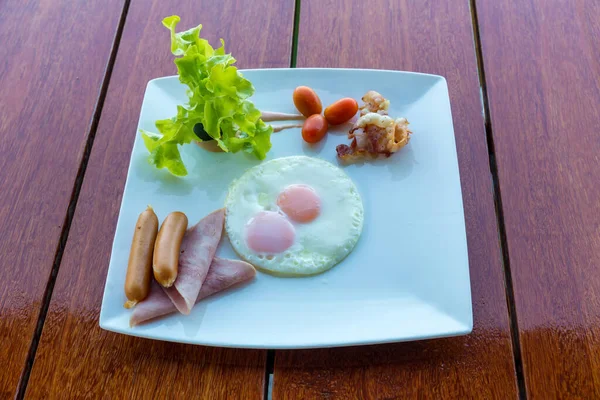 Amerikan kahvaltısı sosisi, yumurta, jambon, pastırma ve sebzeler beyaz tabakta. Kızarmış yumurta ve sosisli geleneksel kahvaltı. Tahta masada kızarmış yumurta, sosis ve jambon..