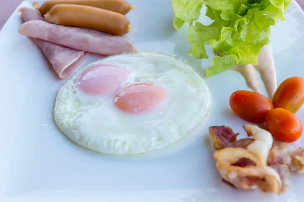 American Breakfast sausage, egg, ham, bacon and vegetables on white plate. Traditional breakfast with fried eggs and sausages. Fried eggs, sausage and ham in breakfast set on wooden table.