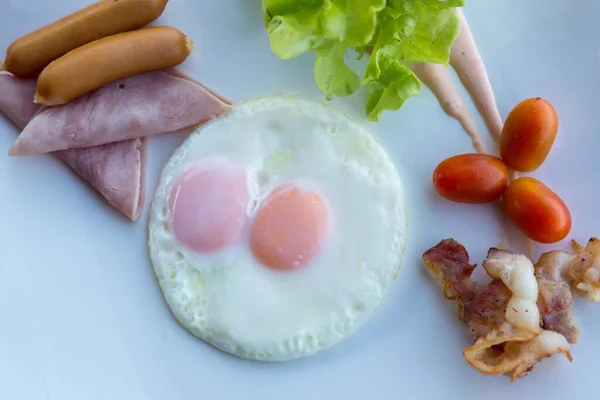 American Breakfast sausage, egg, ham, bacon and vegetables on white plate. Traditional breakfast with fried eggs and sausages. Fried eggs, sausage and ham in breakfast set on wooden table.
