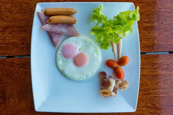 Amerikan kahvaltısı sosisi, yumurta, jambon, pastırma ve sebzeler beyaz tabakta. Kızarmış yumurta ve sosisli geleneksel kahvaltı. Tahta masada kızarmış yumurta, sosis ve jambon..