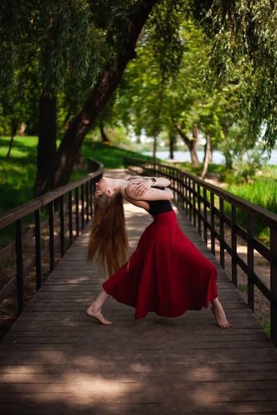 一个年轻的肚皮舞者在公园里的照片 一个年轻的金发姑娘正在大自然中跳舞 穿着红色裙子的女体操运动员 — 图库照片