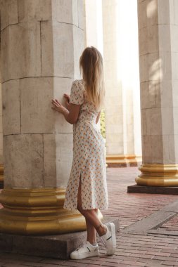 Arka plandaki sütunların arka planında büyüyen bir kızın fotoğrafı. Sarışın bir kız yaz günü şehirde dolaşıyor.