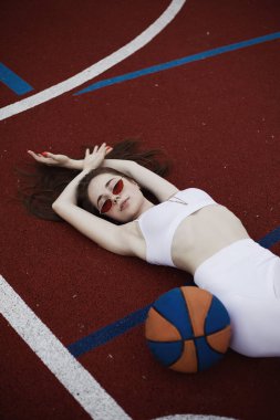 Çekici bir atletik genç bayan sabahları basketbol sahasında yatar, basketbol sahasına yaslanır. Modaya uygun bir fotoğraf. Beyaz spor takım elbiseli esmer kız. Basketbol topu