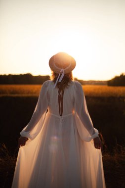 Doğada günbatımında arkası dönük mutlu bir kadın. Kafasında şapkası olan uzun beyaz elbiseli bir kadın.